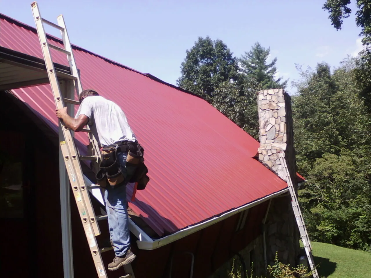 Roofer on a ladder beside a red metal roof and stone chimney — Soffit & Fascia Repair by Roofing Brothers in Stratford